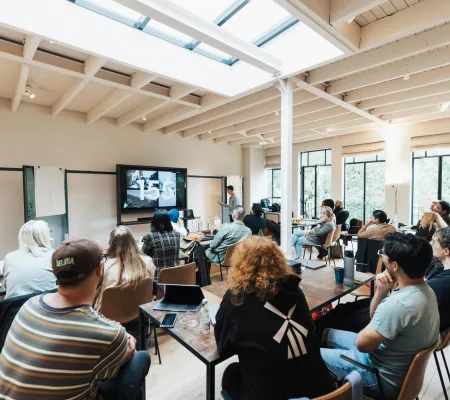 A group seated at tables watching a presentation in a bright meeting room